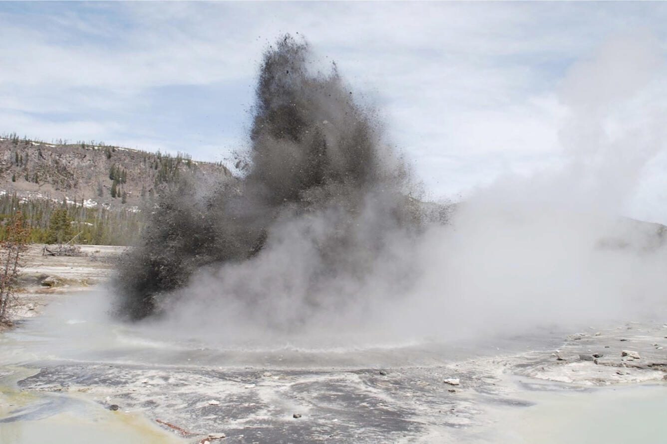 A dark column of rock and steam bursting from the ground at Biscuit Basin
