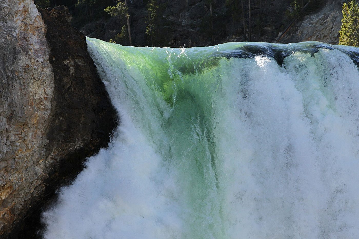 Green water curling over the edge of a waterfall beside a dark rock wall