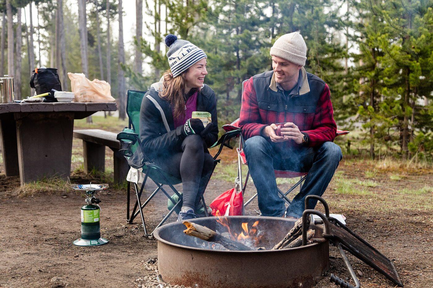 Two campers in hats drinking coffee beside a fire ring on a cold morning at Madison Campground