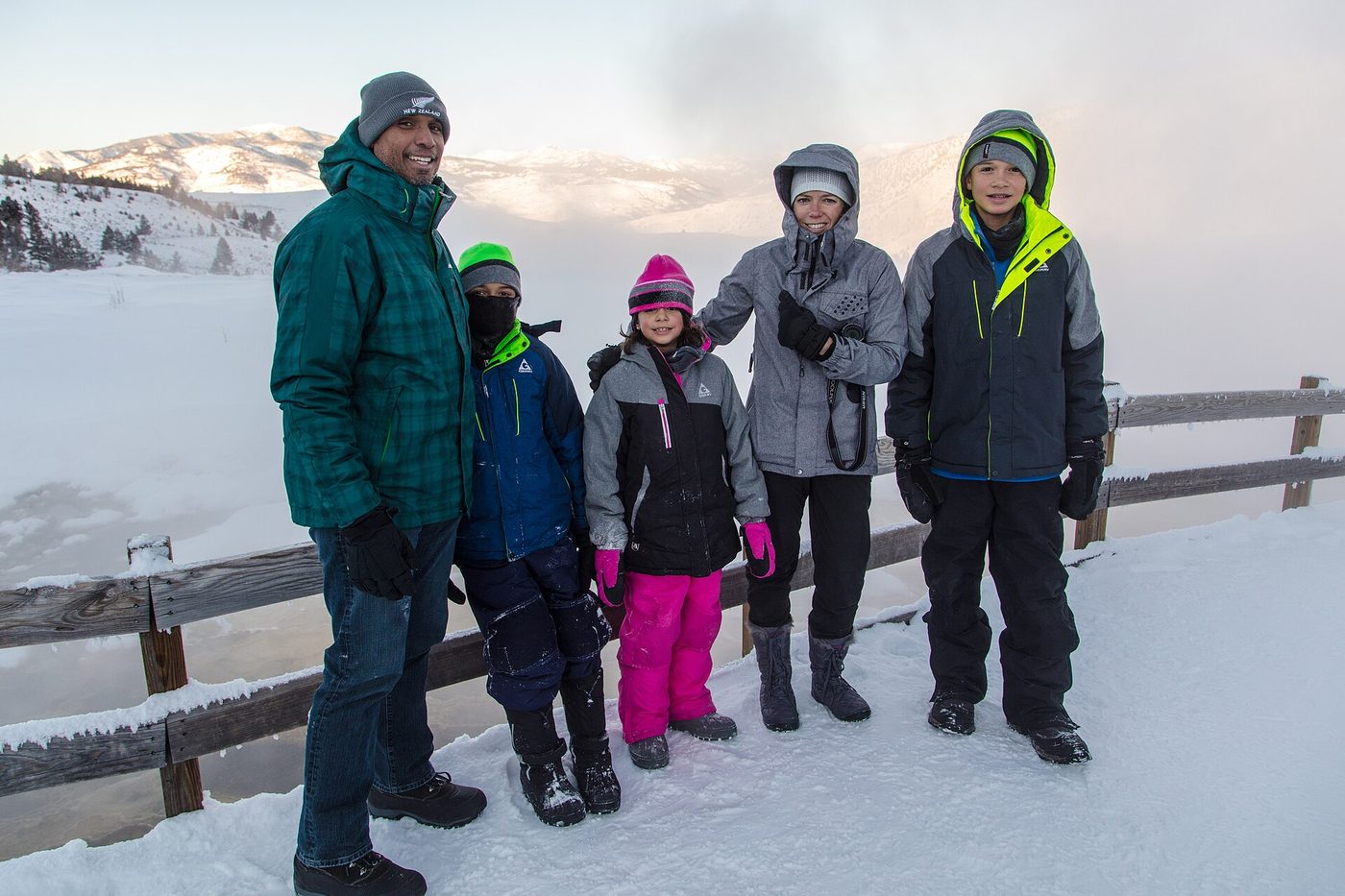 A family in winter coats standing on a snowy boardwalk beside a steaming hot spring