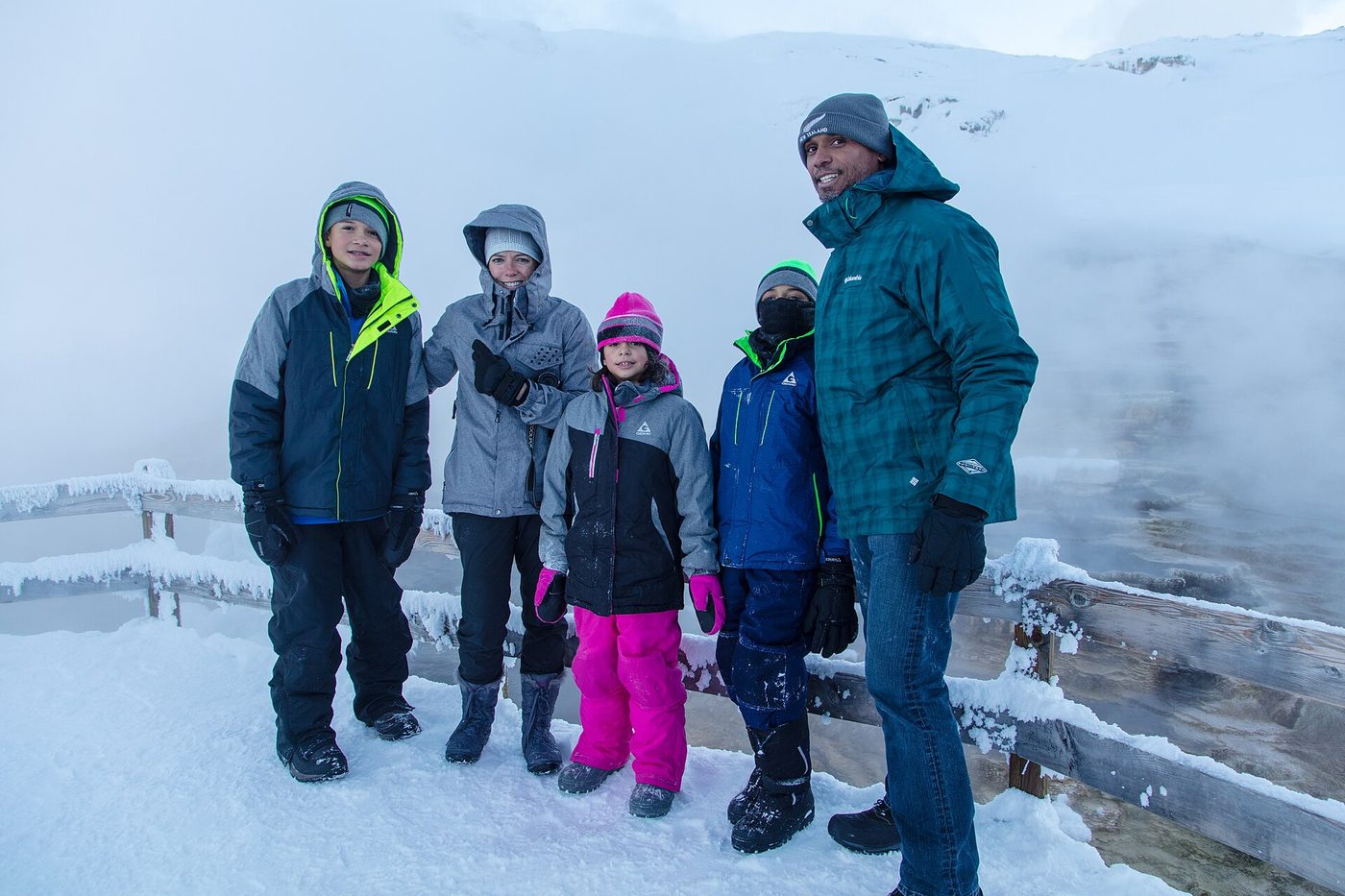 Parents and three children in snow gear standing at a railing above a steaming winter thermal area