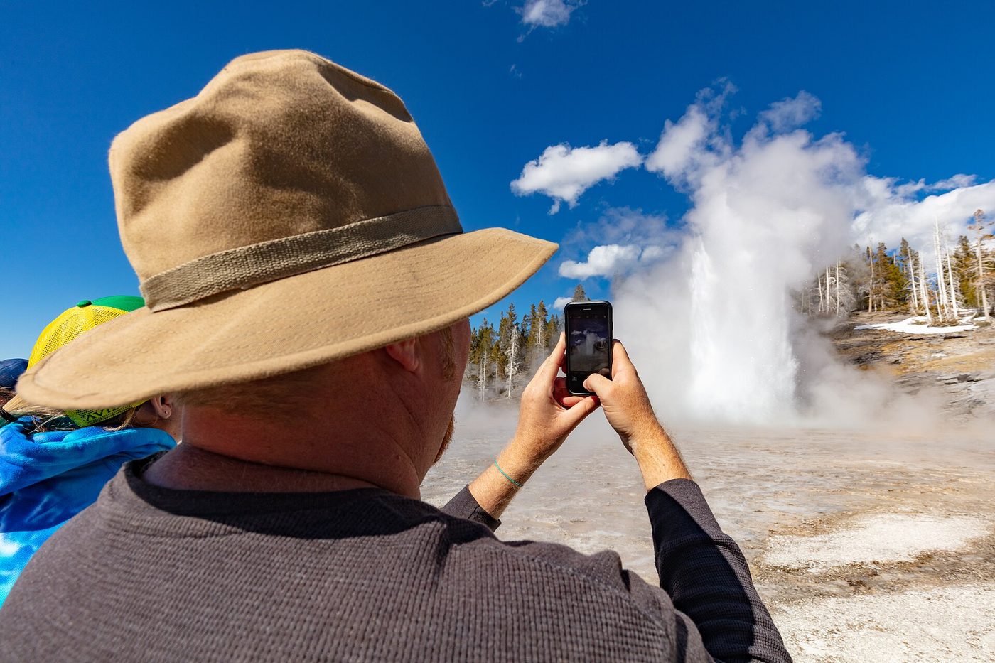Visitor in a sun hat filming a tall geyser eruption on a phone
