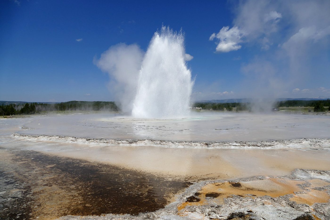 White eruption column rising above wet terraces and a runoff channel under a blue summer sky