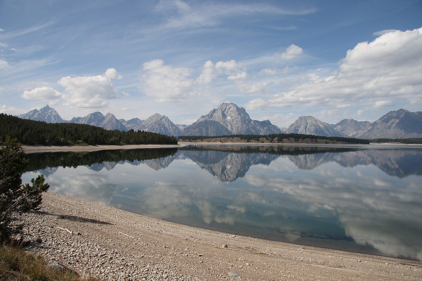 The Teton Range reflected across calm Jackson Lake from a pebble beach