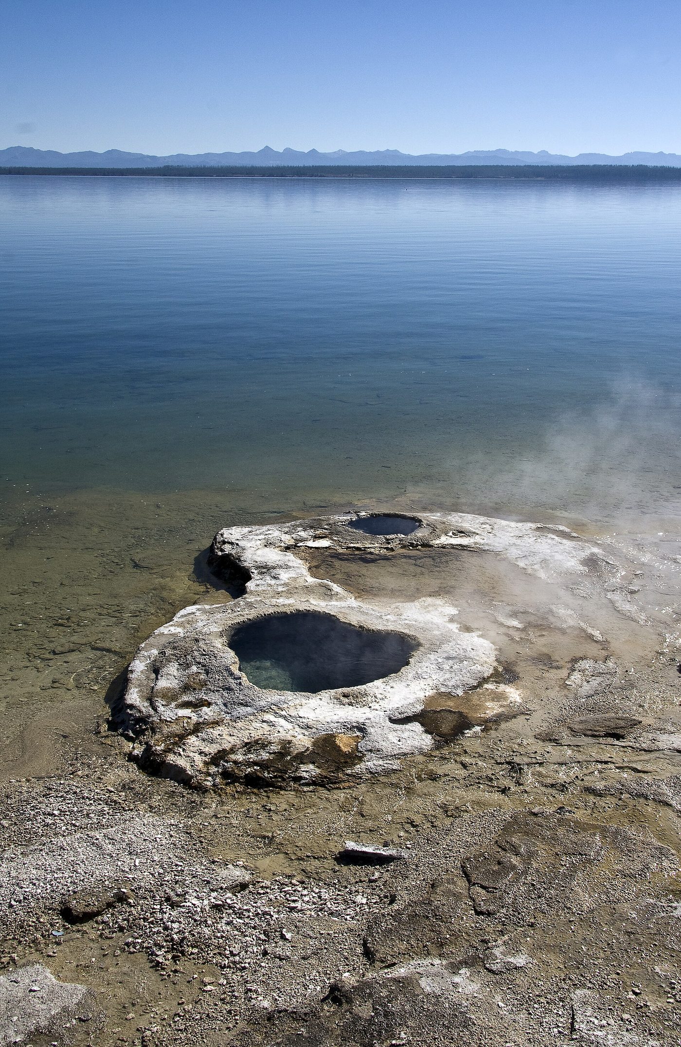 A steaming hot spring cone at the edge of a calm blue lake