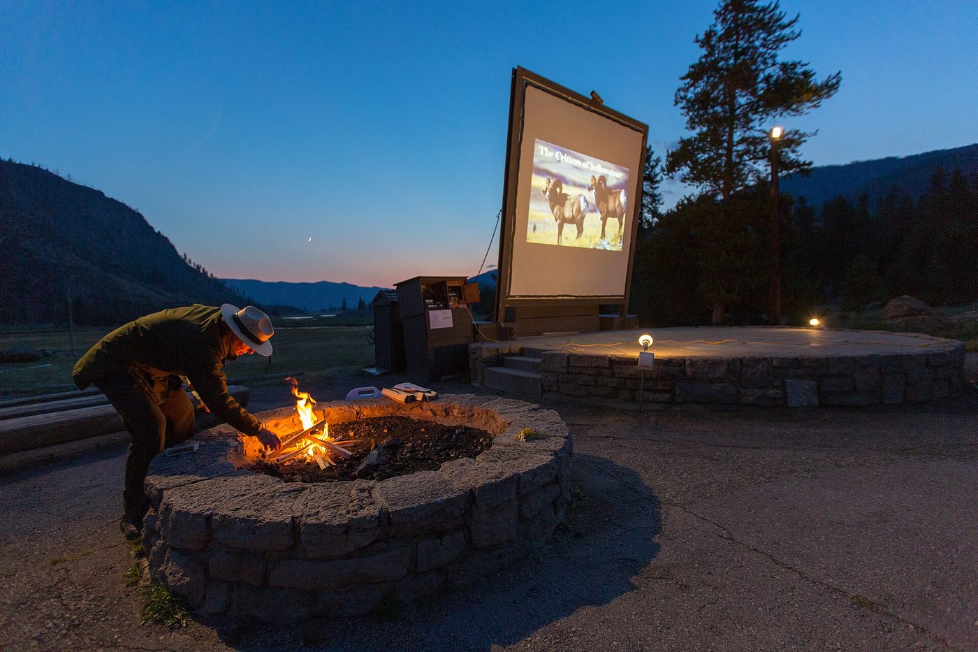 Ranger lighting a campfire in front of the amphitheater screen at dusk