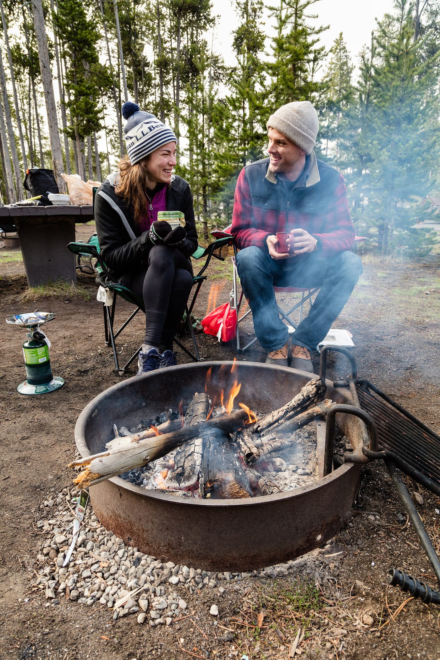 Two campers in warm hats drinking coffee beside a fire ring on a cold morning at Madison