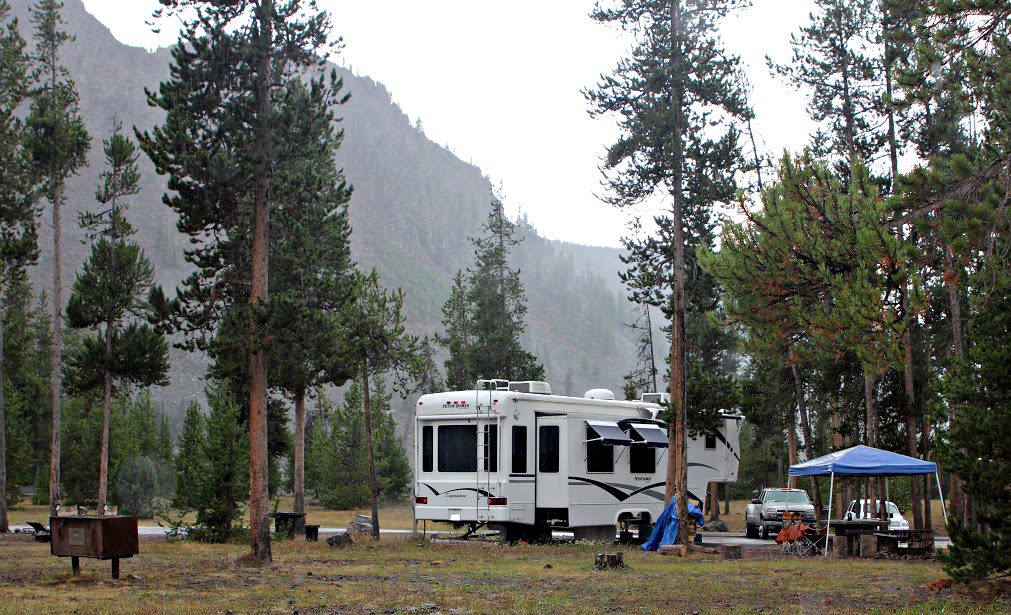 A fifth-wheel trailer parked among lodgepole pines at Madison Campground with a canopy set up nearby