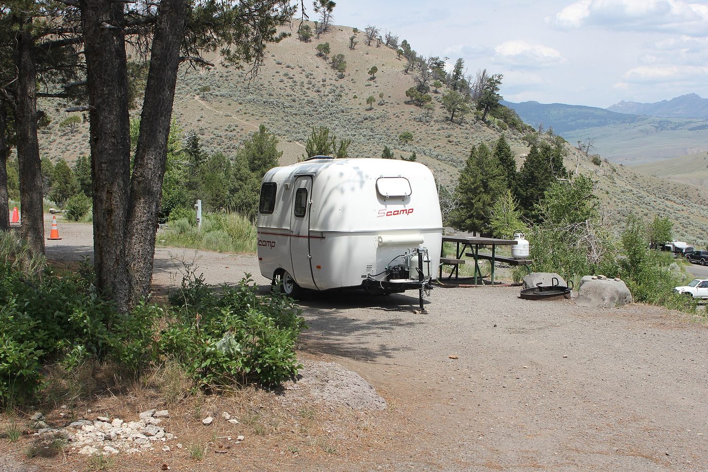 Small fiberglass trailer parked at a sunny sagebrush campsite below dry hills