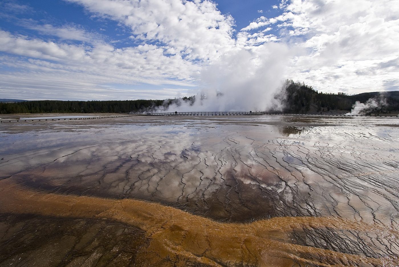 Thin sheets of orange runoff water across white sinter in Midway Geyser Basin, with steam rising beyond