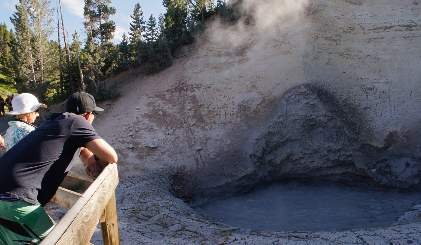 Visitors leaning on a railing above a steaming gray mud pool at the Mud Volcano area
