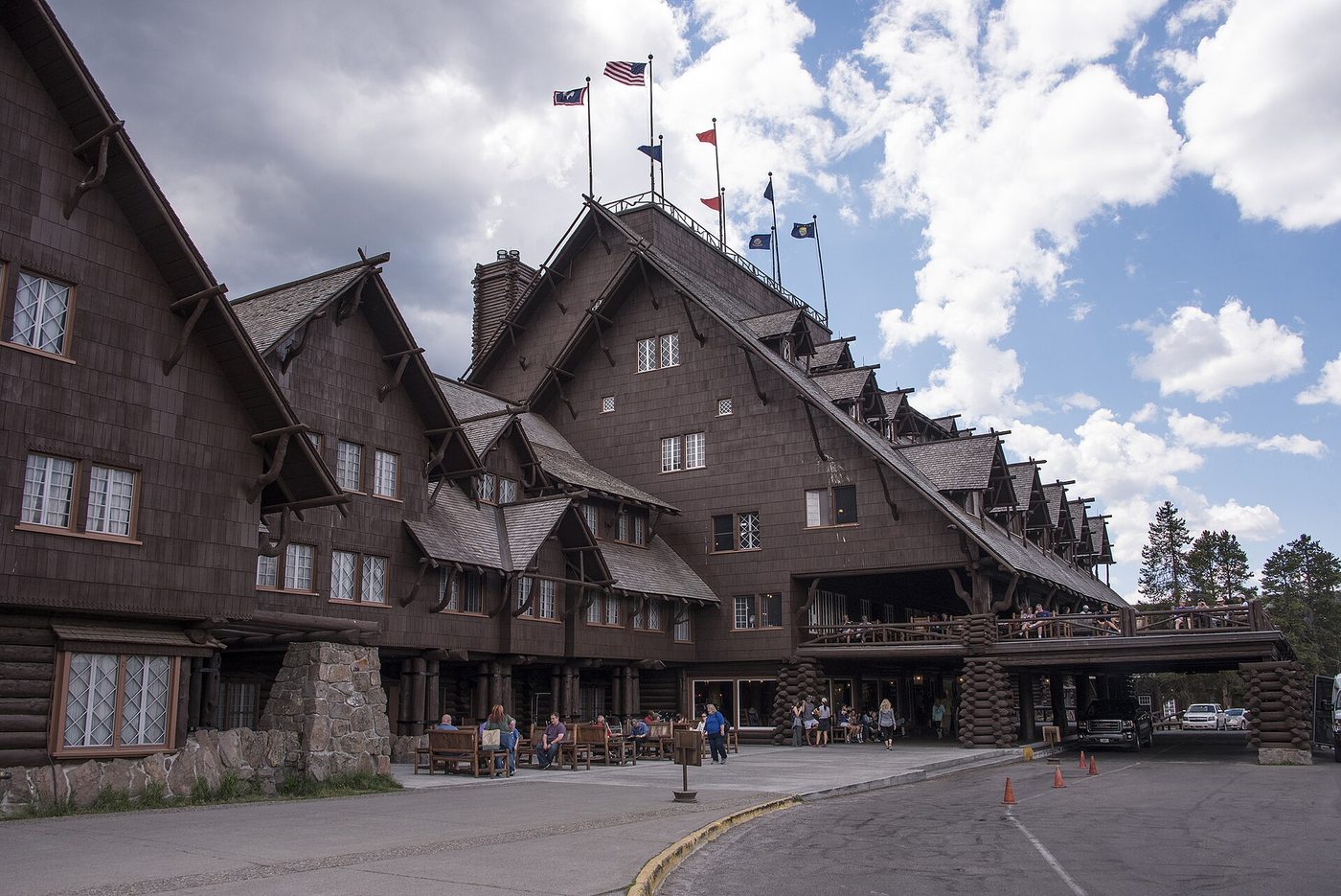 The shingled log facade of the Old Faithful Inn with flags on its roof and visitors on the porch