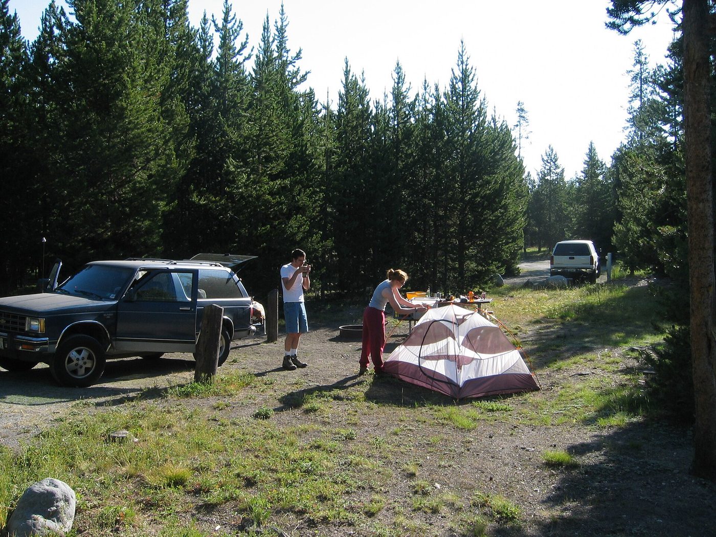 Two campers packing a tent beside their vehicle at a wooded Yellowstone campsite