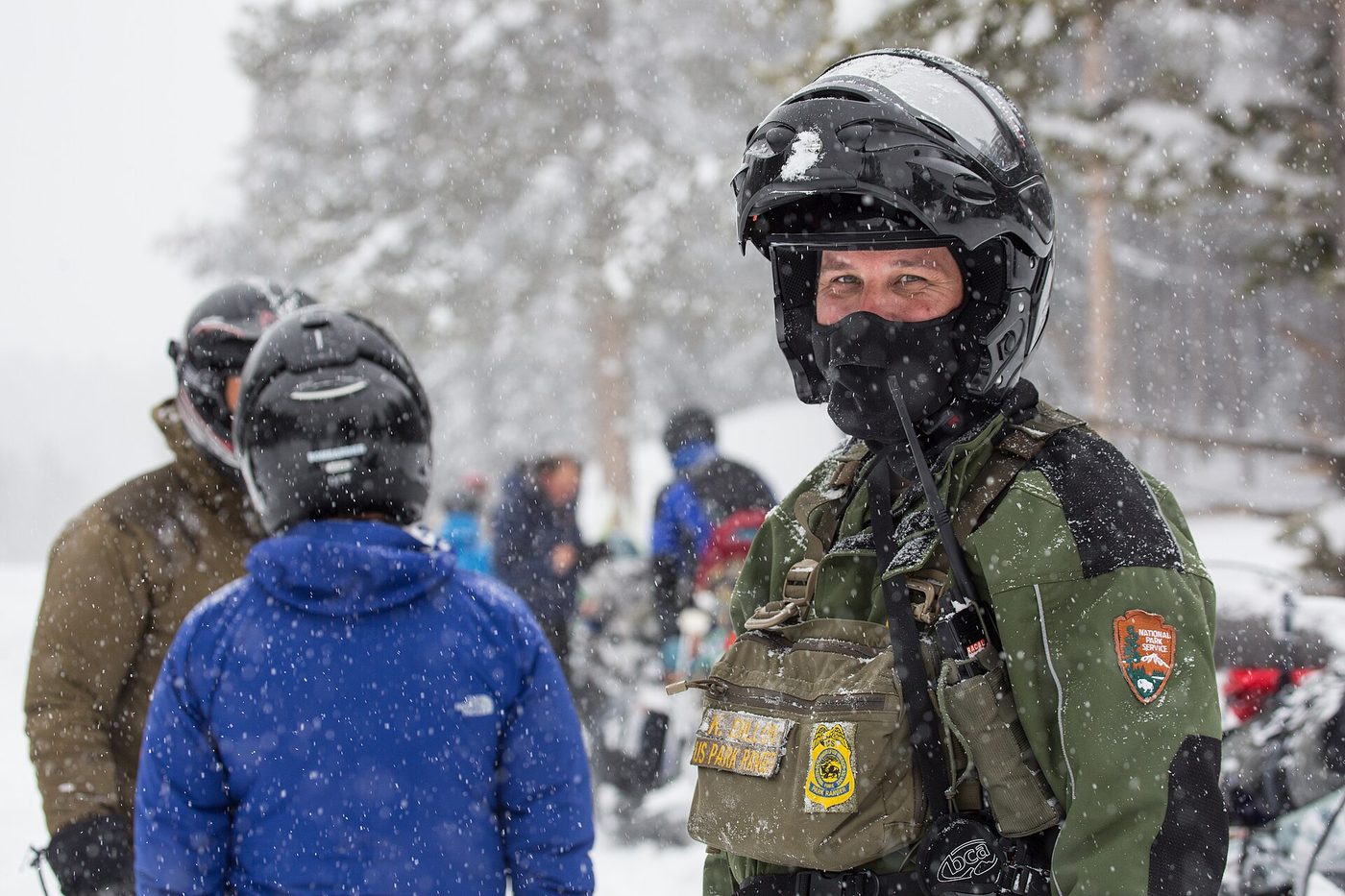 A park ranger in a snowmobile helmet and winter uniform talking with visitors in falling snow