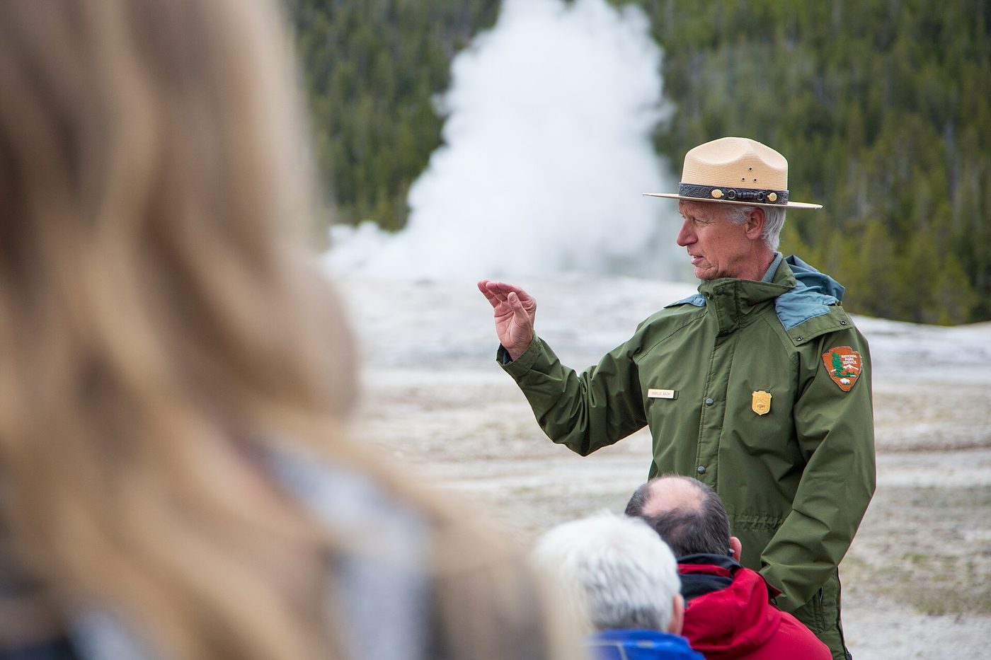 Ranger in a flat hat talking to visitors as a geyser steams in the background