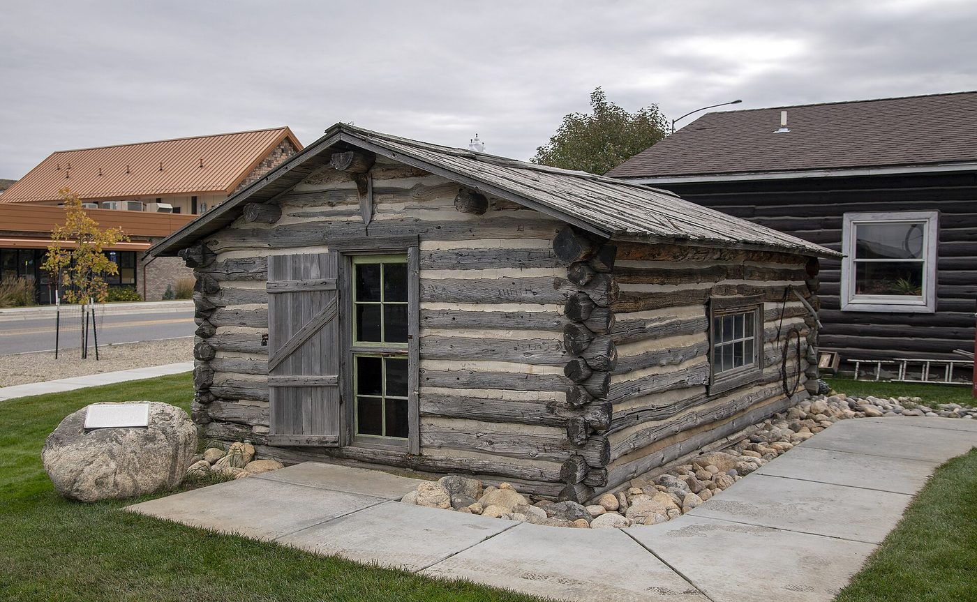 A weathered log homestead cabin with a stone marker on a lawn in Red Lodge, Montana