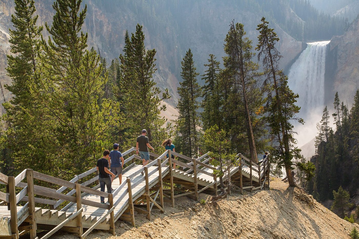 Hikers on wooden stairs descending through pines with a tall waterfall behind