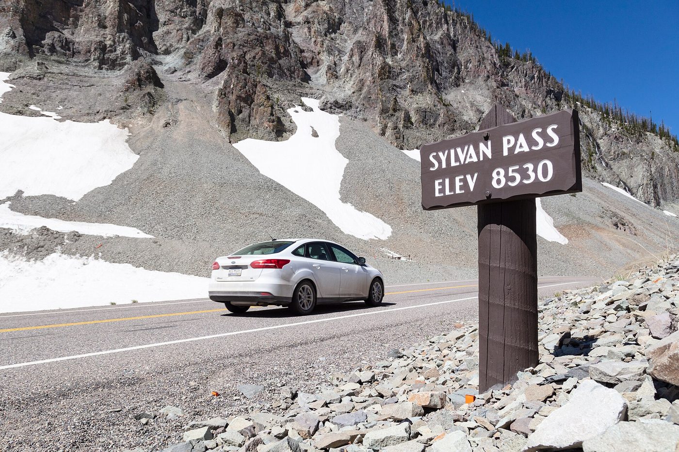 Roadside sign reading Sylvan Pass Elev 8530 with a car passing below scree slopes