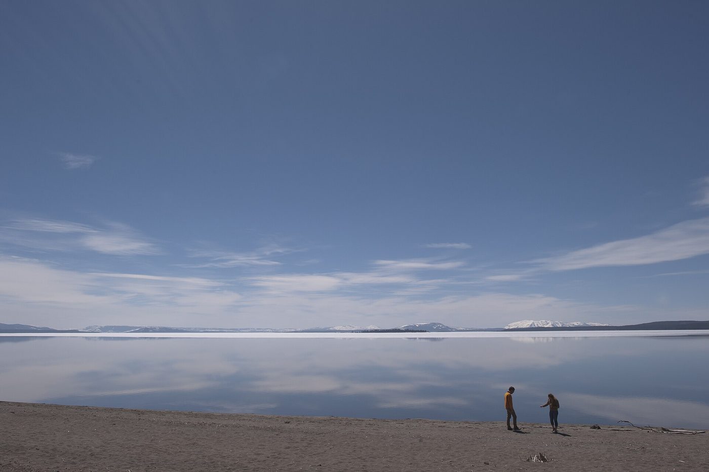 Two visitors on a sandy beach at Yellowstone Lake in May, with ice still covering the far water and snowy peaks beyond